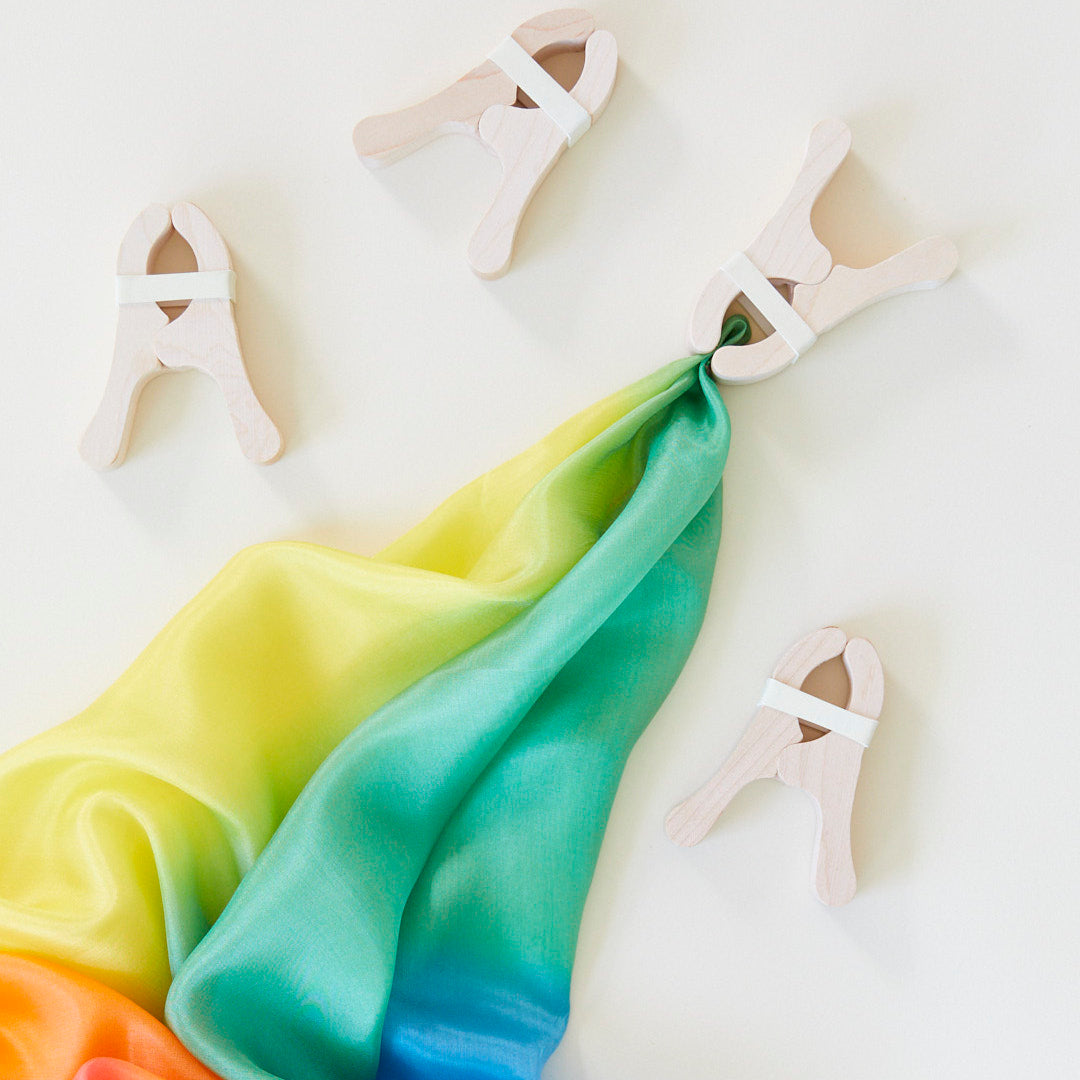 Four wooden clips hold a rainbow silk scarf against a white background, inviting creative play with open-ended toys like Fort Kits.