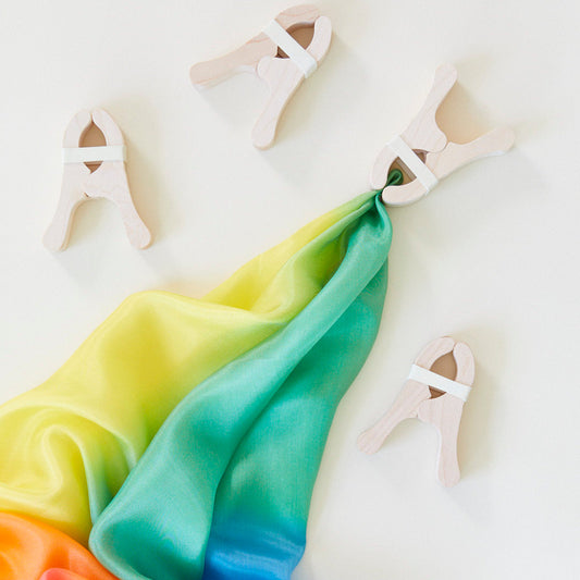 Four wooden clips hold a rainbow silk scarf against a white background, inviting creative play with open-ended toys like Fort Kits.
